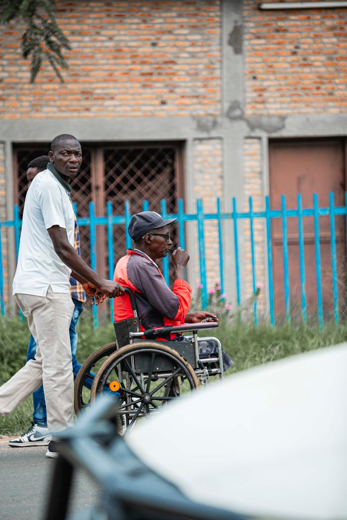 A man assists another in a wheelchair on an urban street.